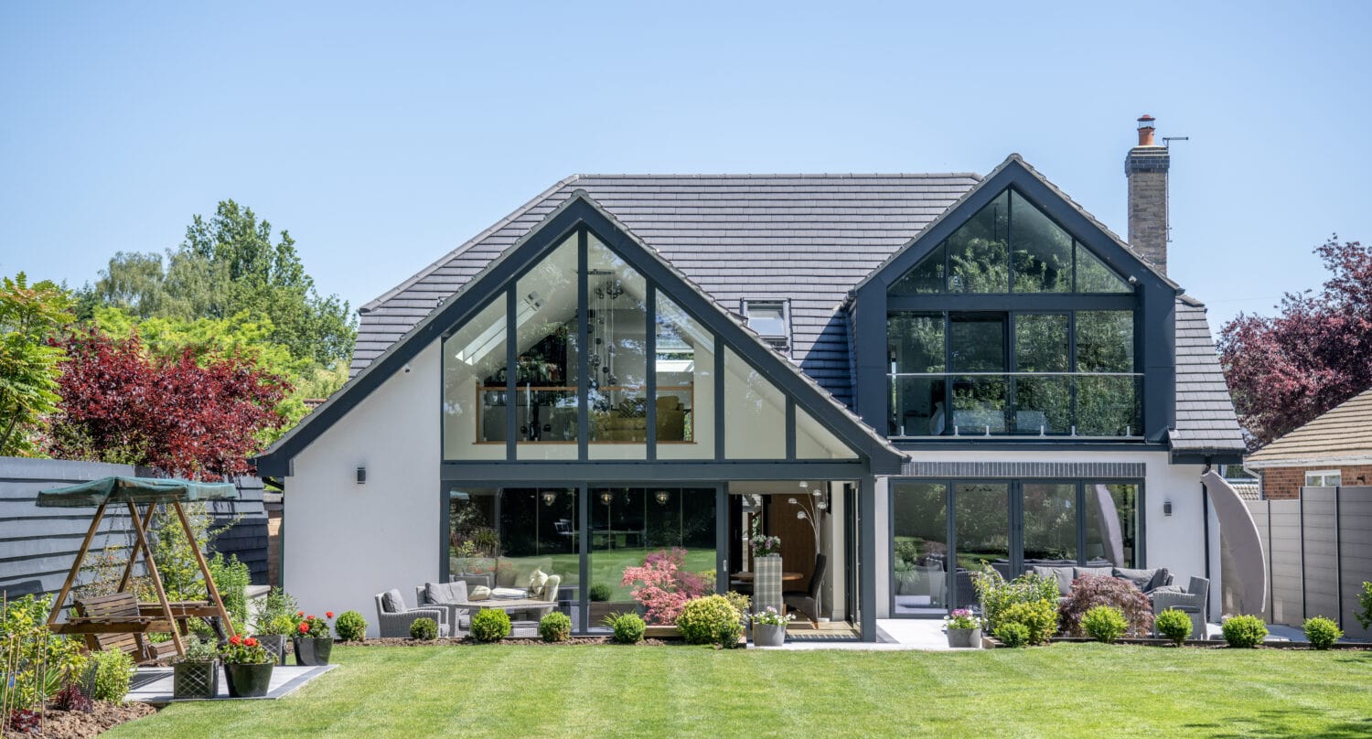 Modern two-story house with large glass windows, bifold doors, gray roof, and white exterior surrounded by a well-kept lawn, colorful shrubs, and trees. A wooden swing and seating area are visible in the sunny backyard.