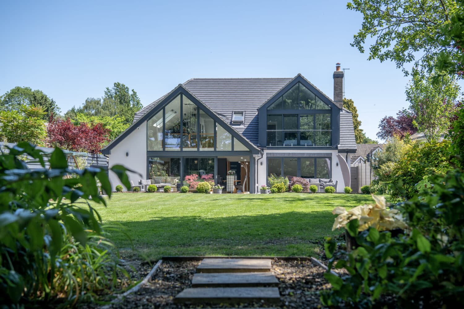 A modern two-story house with large glass windows and a sloped roof features sleek sliding doors, sitting in the center of a lush, green garden with a well-kept lawn, surrounded by trees and shrubs under a clear blue sky.
