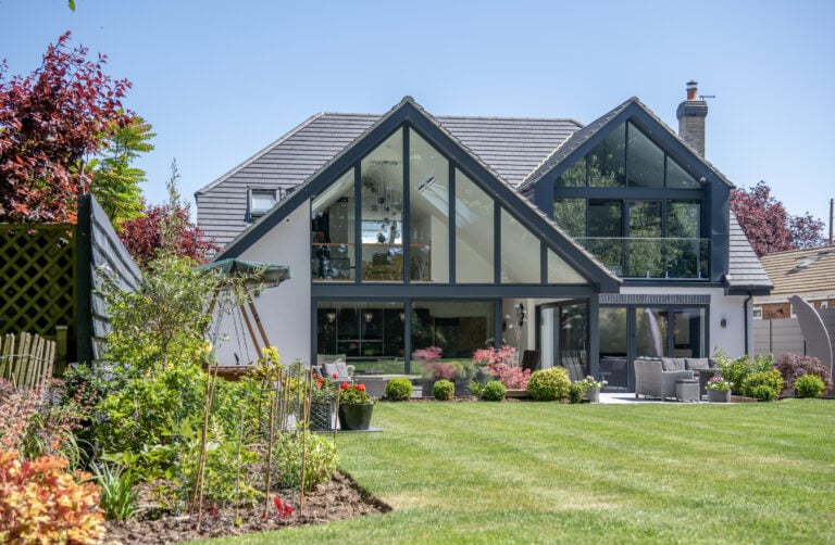 Modern house with large glass windows and a peaked roof, featuring elegant sliding doors opening to a lush green lawn and colorful landscaped garden under a clear blue sky. Outdoor seating area and plants are visible in the foreground.