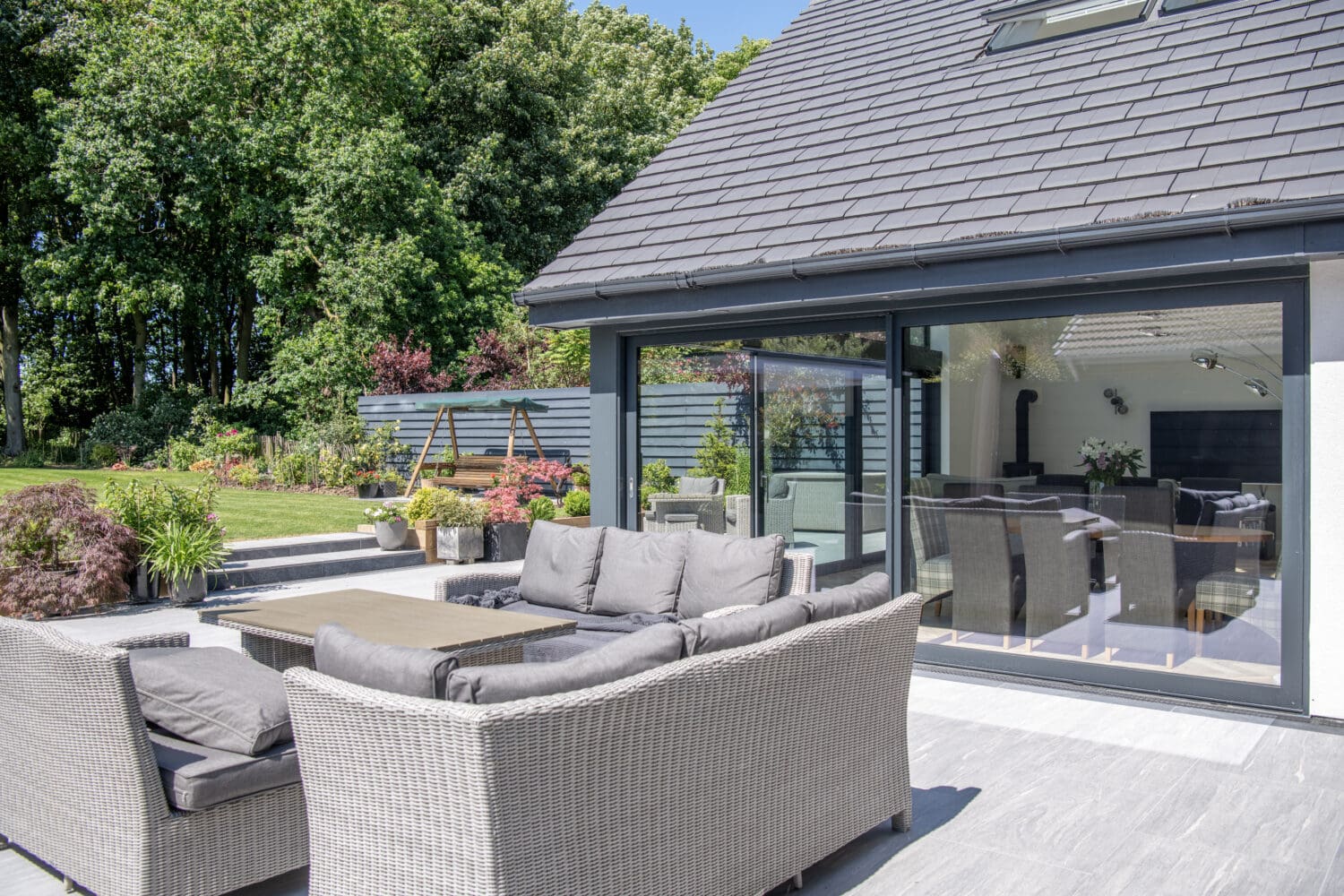 Modern patio with gray wicker sofas and table, set on a tiled terrace beside a house with large sliding doors. The garden features lush greenery, flowers, and a wooden swing beneath trees in the background. Consider adding glass roofing for year-round enjoyment.