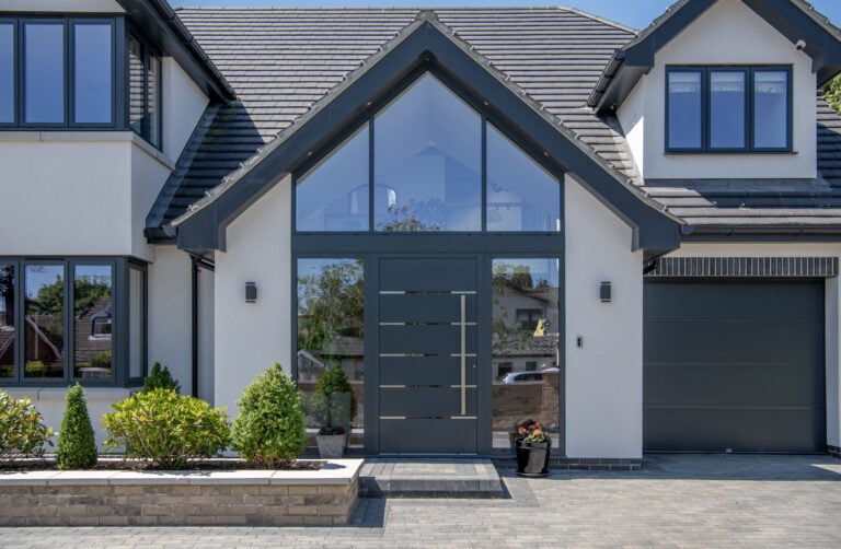 Modern house exterior with bespoke glass solutions, featuring a large glass front door, tall peaked roof, grey trim, symmetrical windows, a driveway, small garden beds with shrubs, and a single-car garage on the right.