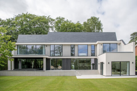 Modern two-story house with large windows, bespoke glass solutions, flat white exterior walls, and a dark sloped roof, surrounded by green grass and trees under a partly cloudy sky.