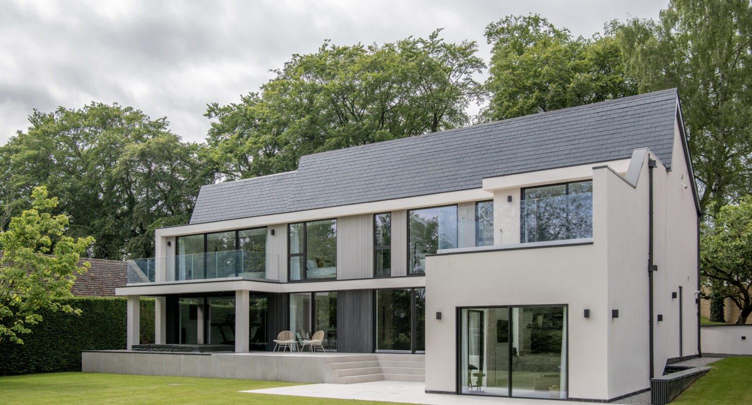 A modern two-story house with large glass windows, bespoke glass solutions like bifold doors, a balcony, and a spacious patio, surrounded by a green lawn and tall trees under a cloudy sky.