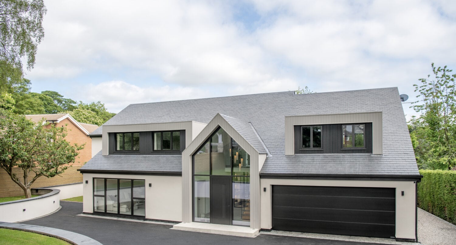 Modern two-story house with large windows, a gray gabled roof, black garage doors, and bespoke glass solutions at the entryway. The home is surrounded by a curved driveway, trees, and greenery under a partly cloudy sky.