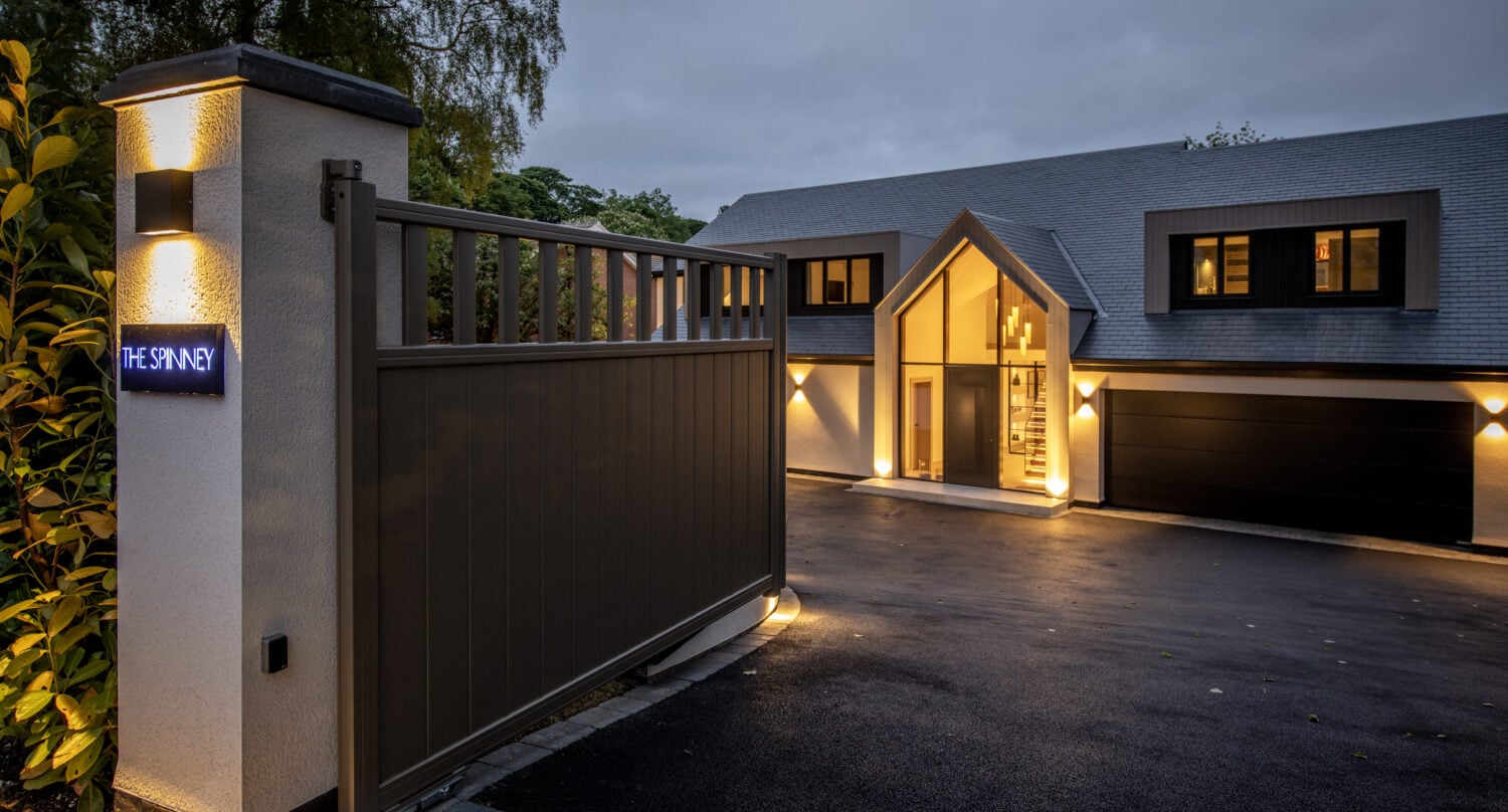 Modern house with large windows, bifold doors, and a sloped roof illuminated at dusk. A tall gate with a sign reading THE SPINNEY is open at the entrance, leading to a driveway and double garage. Trees are visible in the background.