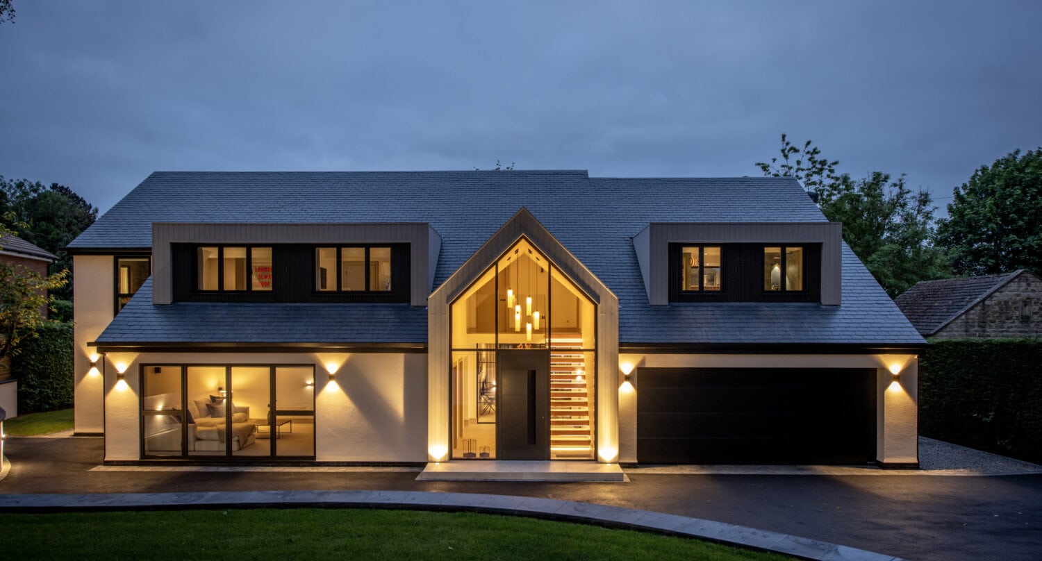Modern two-story house at dusk with large windows, black and white exterior, bespoke glass solutions on the illuminated entrance with a glass front, visible staircase, and a double garage, surrounded by greenery and a dark driveway.