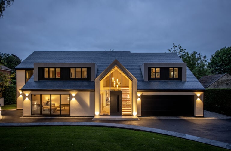 Modern two-story house at dusk with large windows, black and white exterior, bespoke glass solutions on the illuminated entrance with a glass front, visible staircase, and a double garage, surrounded by greenery and a dark driveway.