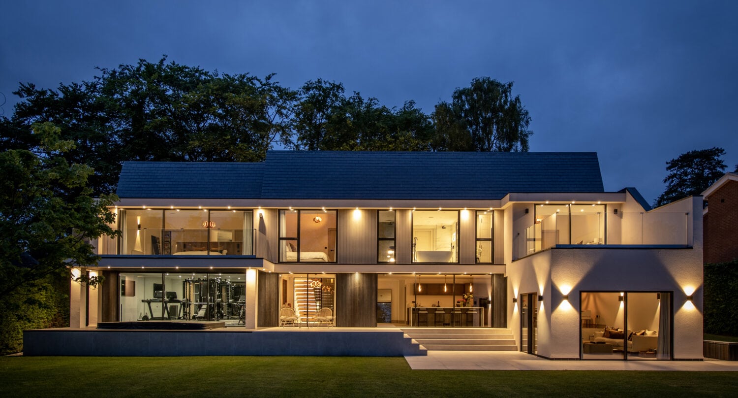 Modern two-story house at dusk with large glass windows, bespoke glass solutions like bifold doors and glass roofing, illuminated interior and exterior lights, a visible home gym, open living spaces, and a well-manicured lawn in the foreground.