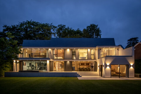 Modern two-story house at dusk with large glass windows, bespoke glass solutions like bifold doors and glass roofing, illuminated interior and exterior lights, a visible home gym, open living spaces, and a well-manicured lawn in the foreground.