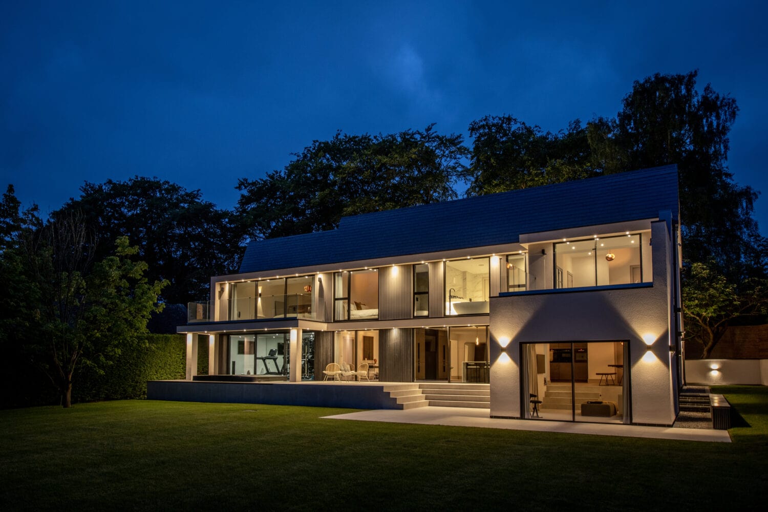 A modern two-story house with large windows and bright interior lights, viewed at dusk. Sliding doors open to a well-kept lawn, while bespoke glass solutions enhance the homes sleek, contemporary design amid surrounding trees.