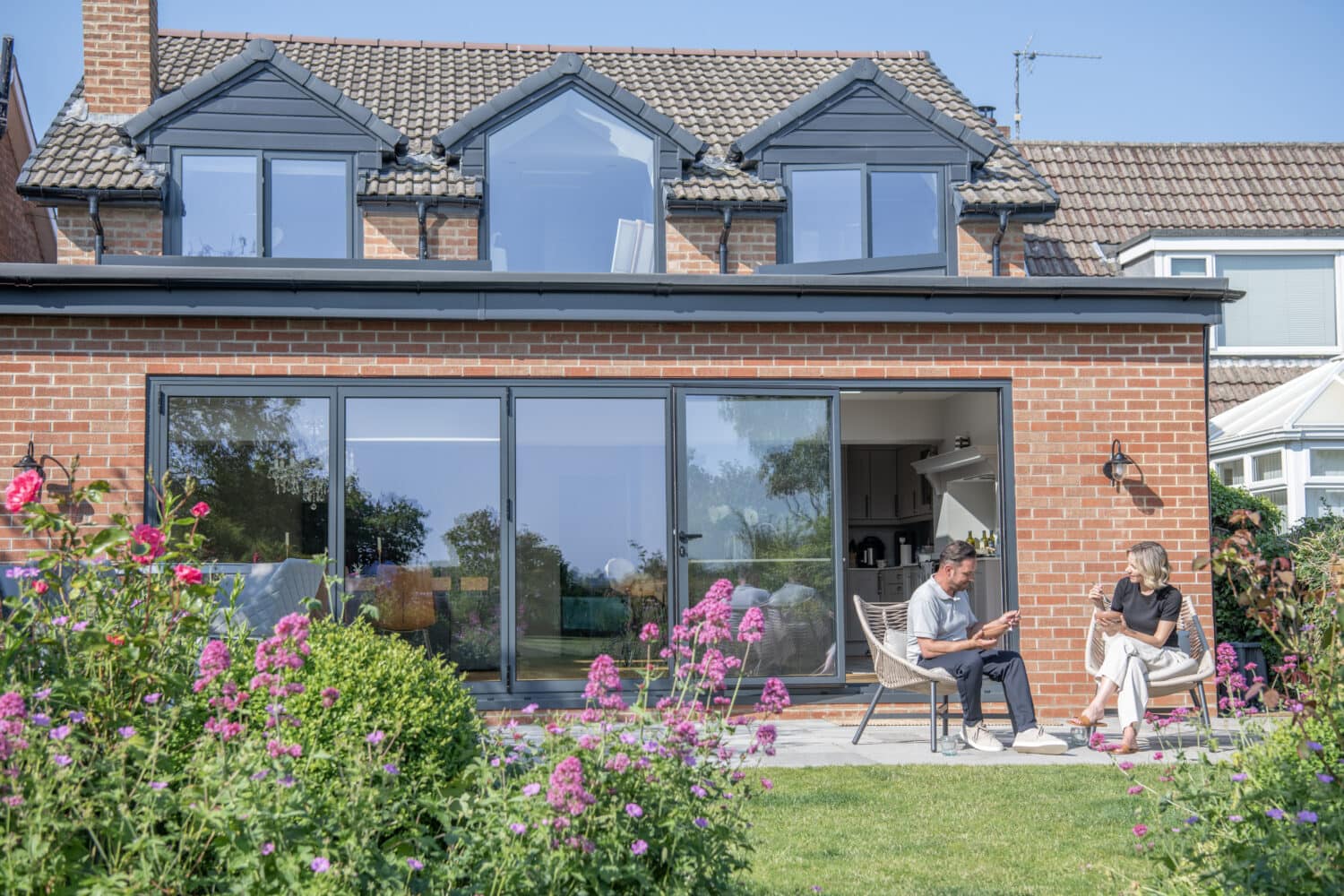 A man and woman sit talking on a patio outside a modern brick house with large sliding glass bifold doors, surrounded by a garden with pink and green plants on a sunny day.