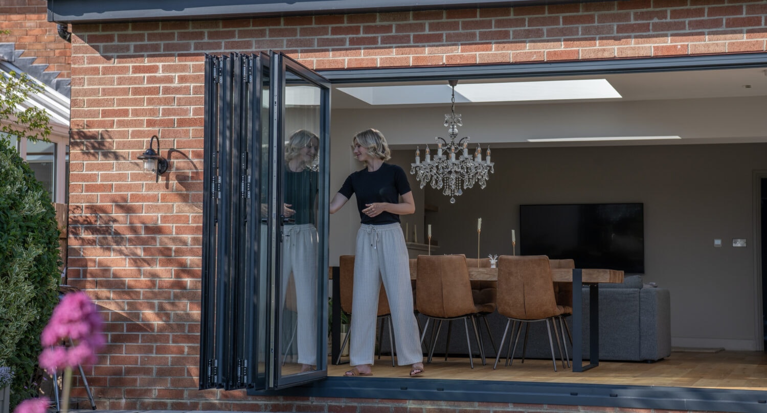 A woman stands by open bifold doors of a modern brick house, looking outside. Inside, there’s a dining table with brown chairs, a chandelier, and a couch. Pink flowers are visible in the foreground.