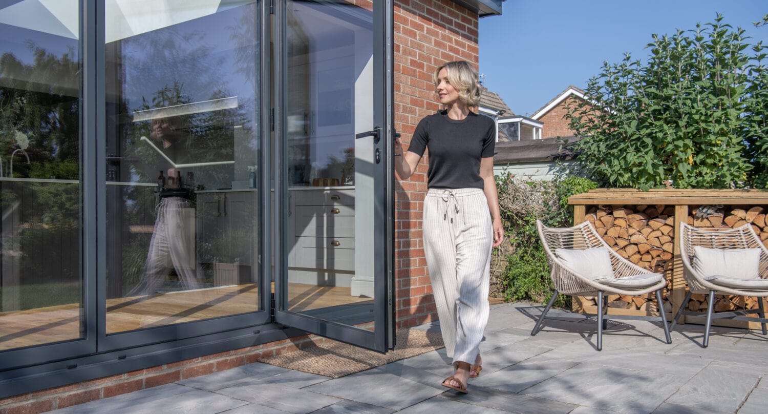 A woman in a black t-shirt and light pants walks barefoot from modern bifold doors onto a stone patio, with outdoor chairs and a stacked woodpile in the background on a sunny day.