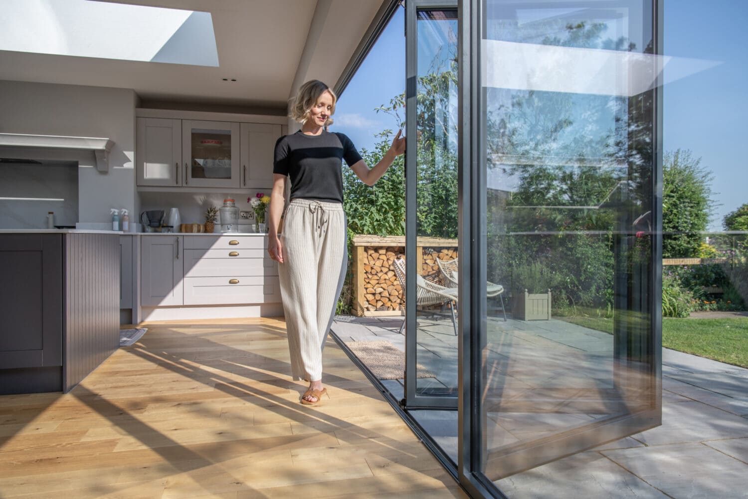 A woman in casual clothes opens large bifold doors from a modern kitchen onto a sunny patio and garden, letting in natural light.