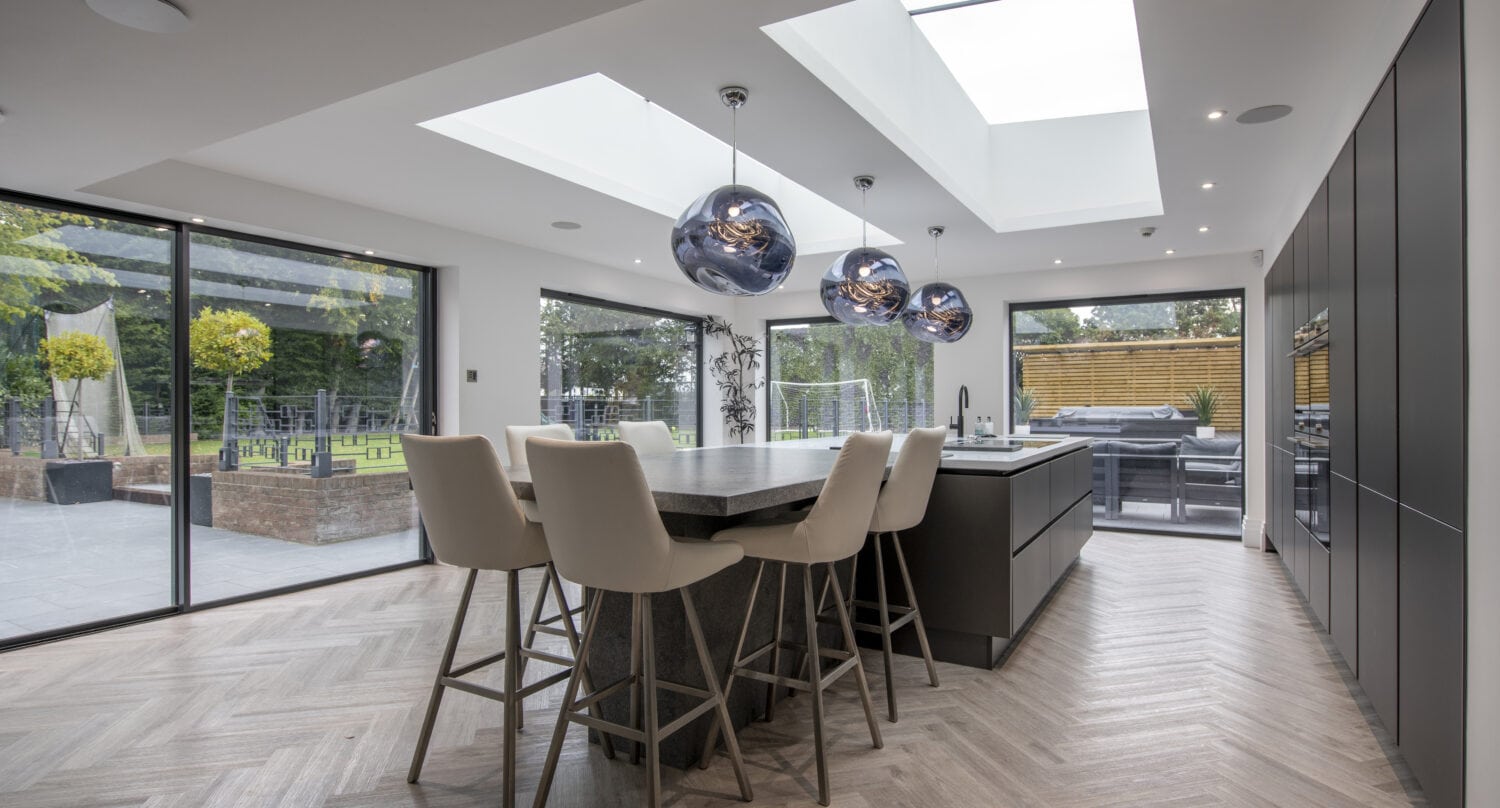 Modern kitchen with large island, four barstools, and three pendant lights. Skylights and floor-to-ceiling sliding doors fill the space with natural light. Outdoor patio and garden are visible through the expansive sliding glass doors.