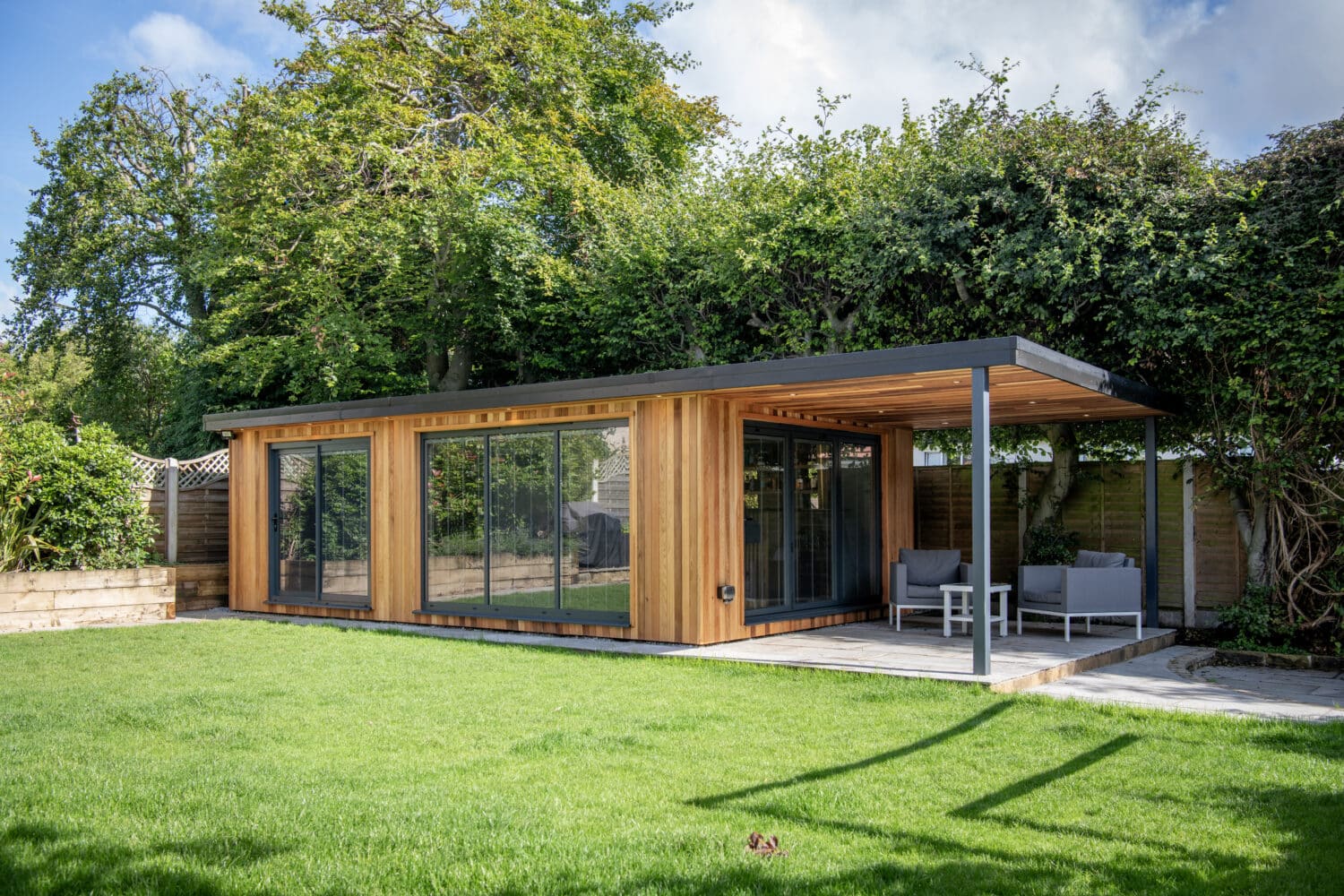 A modern garden room with wooden siding and large glass windows features bifold doors opening onto a covered patio with outdoor seating. Trees and shrubs surround the structure, providing privacy, greenery, and a seamless indoor-outdoor flow.