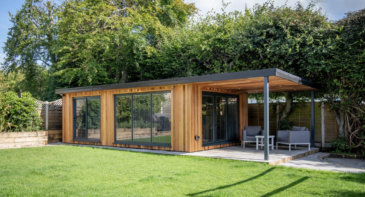 A modern garden room with wooden siding and large glass windows features bifold doors opening onto a covered patio with outdoor seating. Trees and shrubs surround the structure, providing privacy, greenery, and a seamless indoor-outdoor flow.