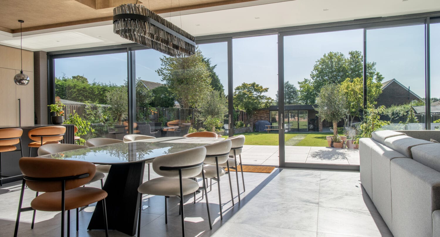 Modern dining and living area with a glass table, mixed color chairs, and a gray sofa, featuring large floor-to-ceiling sliding doors overlooking a sunny garden with green trees and patio plants.