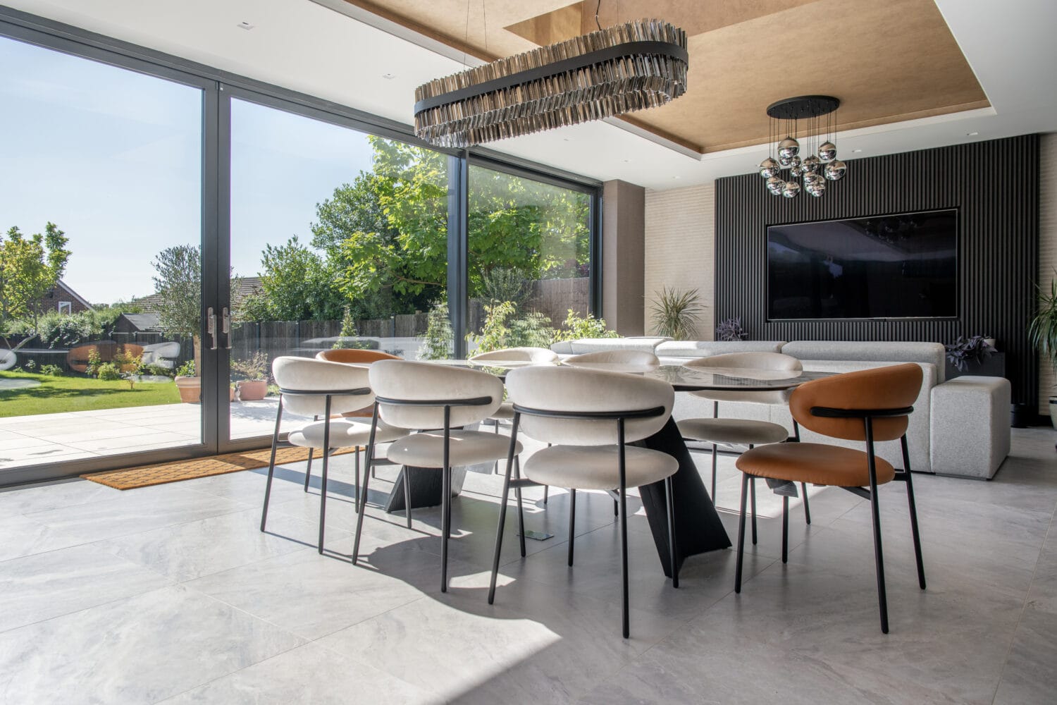 Modern dining area with a long table and mixed-color chairs, bifold doors opening to a sunny garden, grey sofa, wall-mounted TV, and unique ceiling lights in a bright, spacious room.