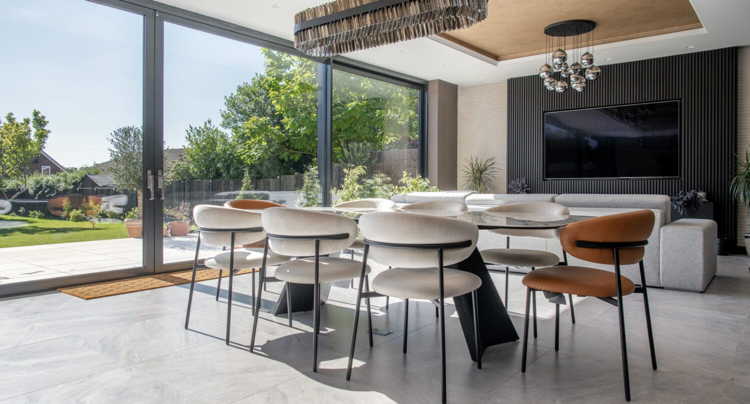 Modern dining area with a long table and mixed-color chairs, bifold doors opening to a sunny garden, grey sofa, wall-mounted TV, and unique ceiling lights in a bright, spacious room.