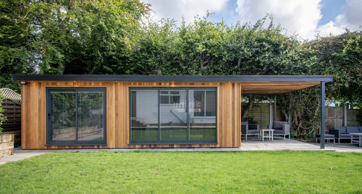A modern garden room with wooden paneling, large glass sliding doors, and a covered patio area featuring bespoke glass solutions, set in a lush green backyard with trees and hedge behind.