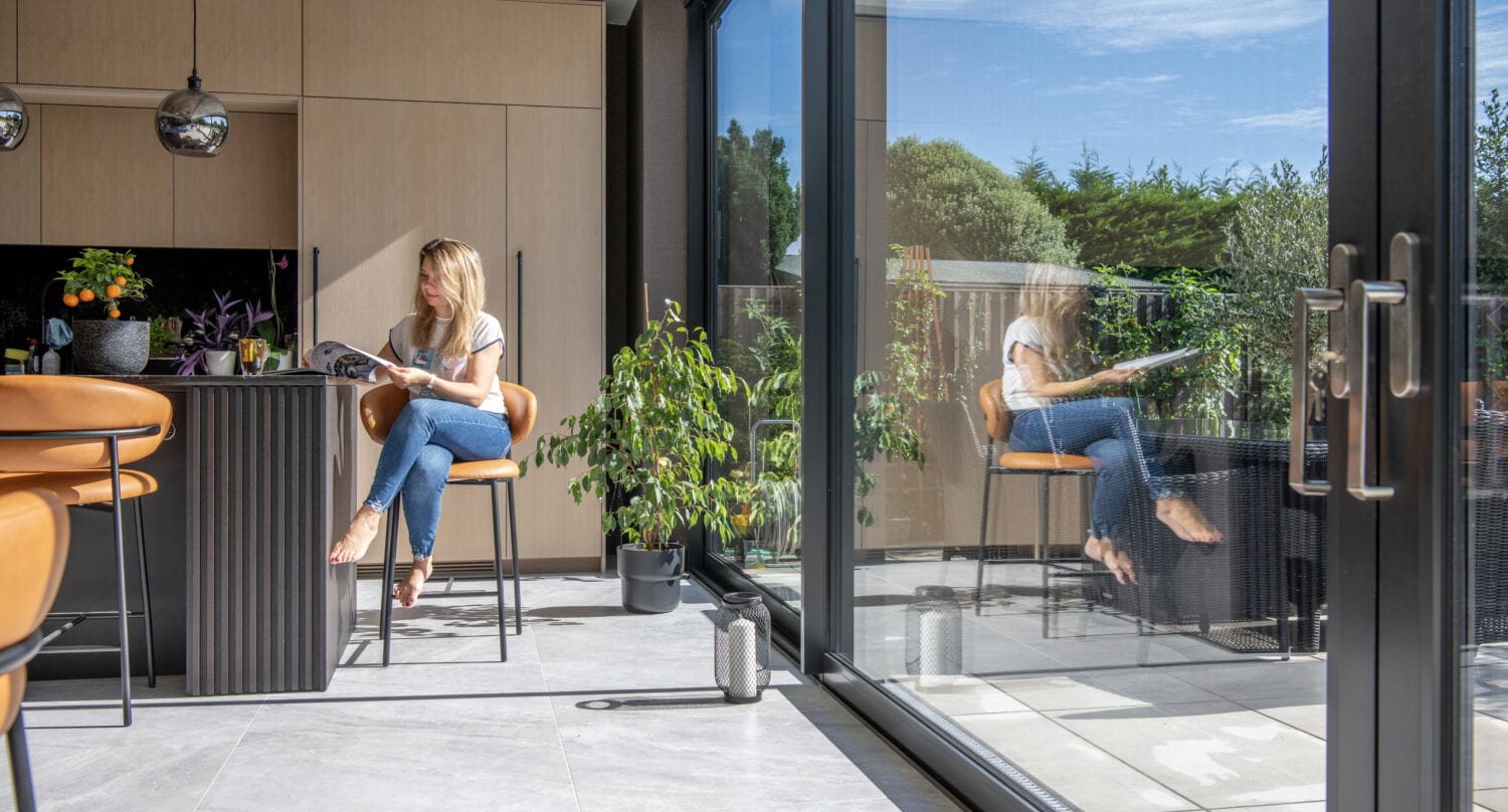 A woman sits on a stool in a modern kitchen, reading a magazine. Bespoke glass solutions create large doors that reflect her image, revealing a sunny patio and green garden. The space is bright with natural light and contemporary decor.