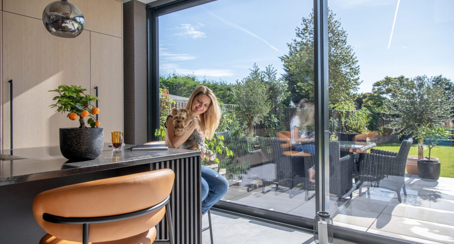 A woman sits by a large glass window in a modern kitchen, holding a small dog. Sunlight streams in through bespoke glass solutions, revealing a lush garden and patio with outdoor furniture. An orange chair and potted plant are in the foreground.