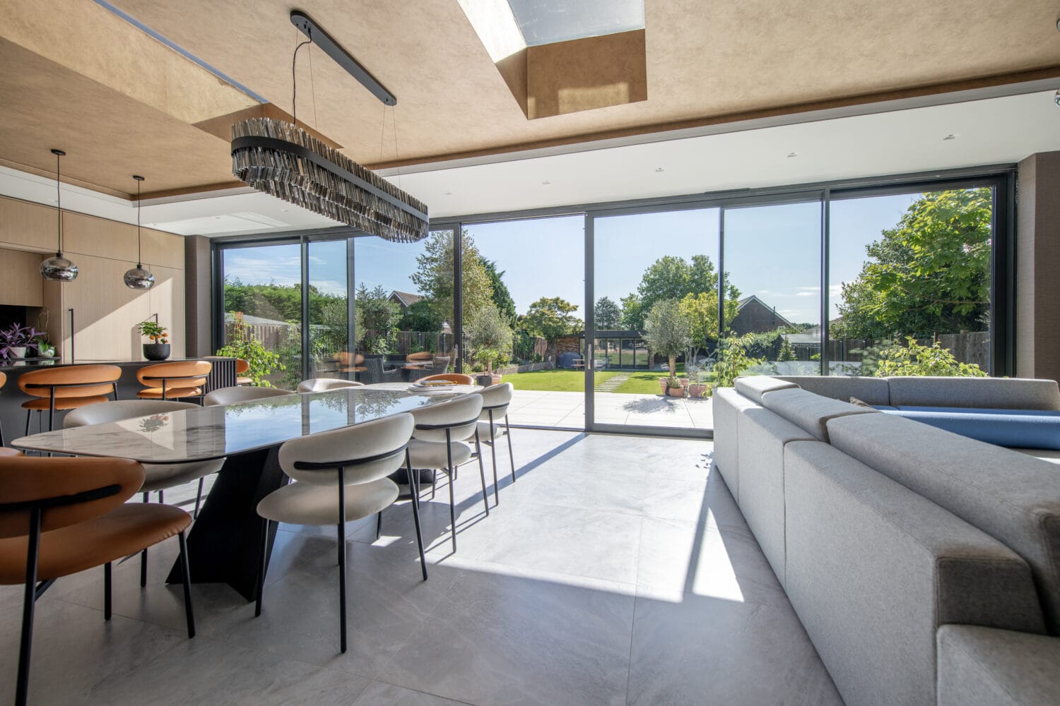 Modern dining and living area with bespoke glass solutions, including large sliding doors opening to a sunny garden. The room features a long dining table with chairs, a contemporary chandelier, kitchen island, and light gray sectional sofa.