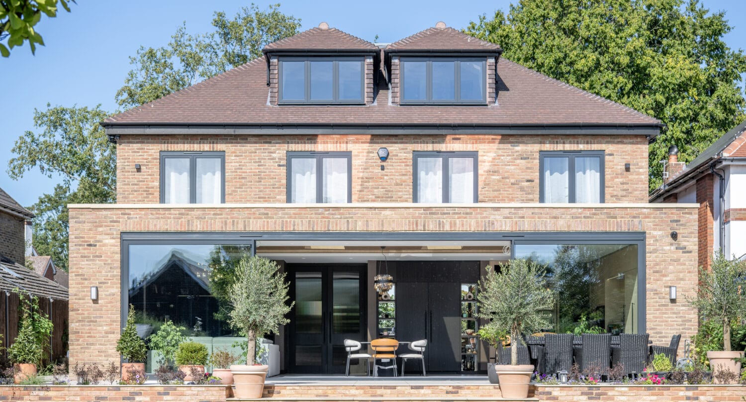 A modern two-story brick house with large windows, sleek glass roofing, a glass sliding door, potted plants, outdoor dining furniture, and a well-kept lawn in the foreground.