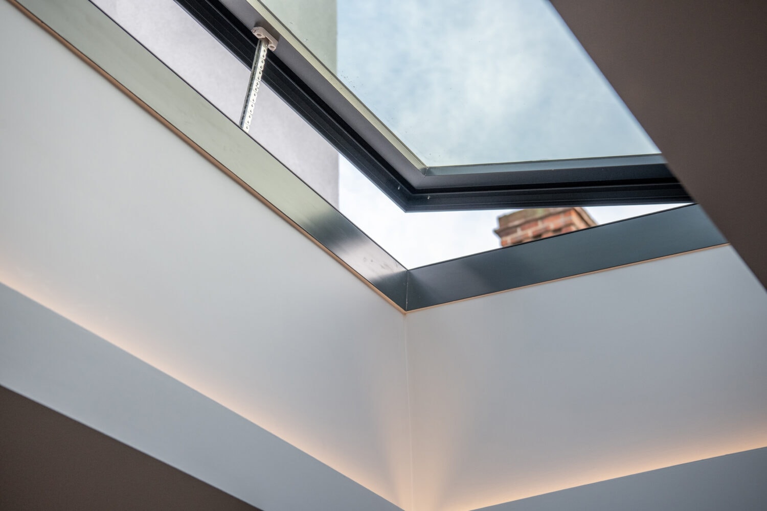 A view looking up at an open skylight window in a modern ceiling with clean lines and soft recessed lighting, featuring bespoke glass solutions that reveal a glimpse of blue sky and part of a building outside.
