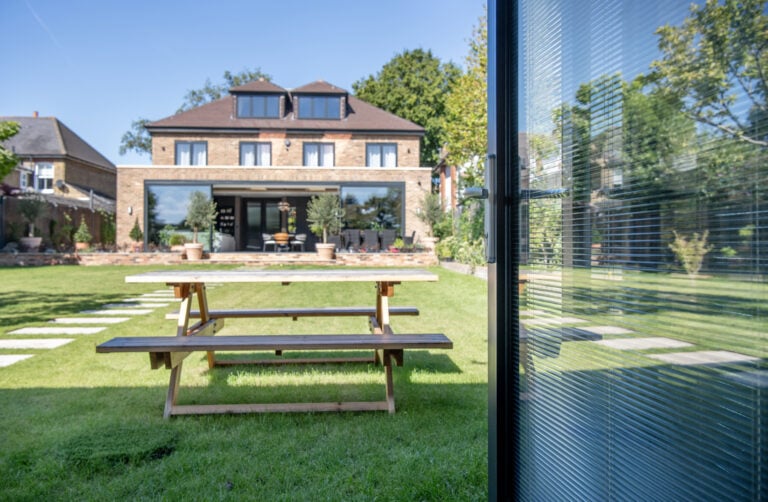 A wooden picnic table sits on a green lawn in a backyard, with a modern brick house and large bifold doors in the background. A glass structure is visible on the right side of the image.