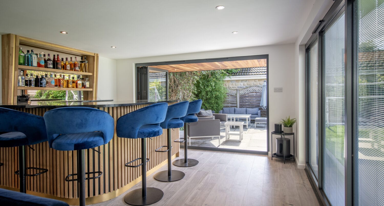 Modern home bar with blue velvet stools, a wooden counter, and a well-stocked shelf of drinks. Bespoke glass solutions with large bifold doors open to a sunny patio with outdoor seating and greenery.