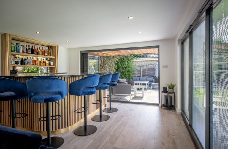 Modern home bar with blue velvet stools, a wooden counter, and a well-stocked shelf of drinks. Bespoke glass solutions with large bifold doors open to a sunny patio with outdoor seating and greenery.