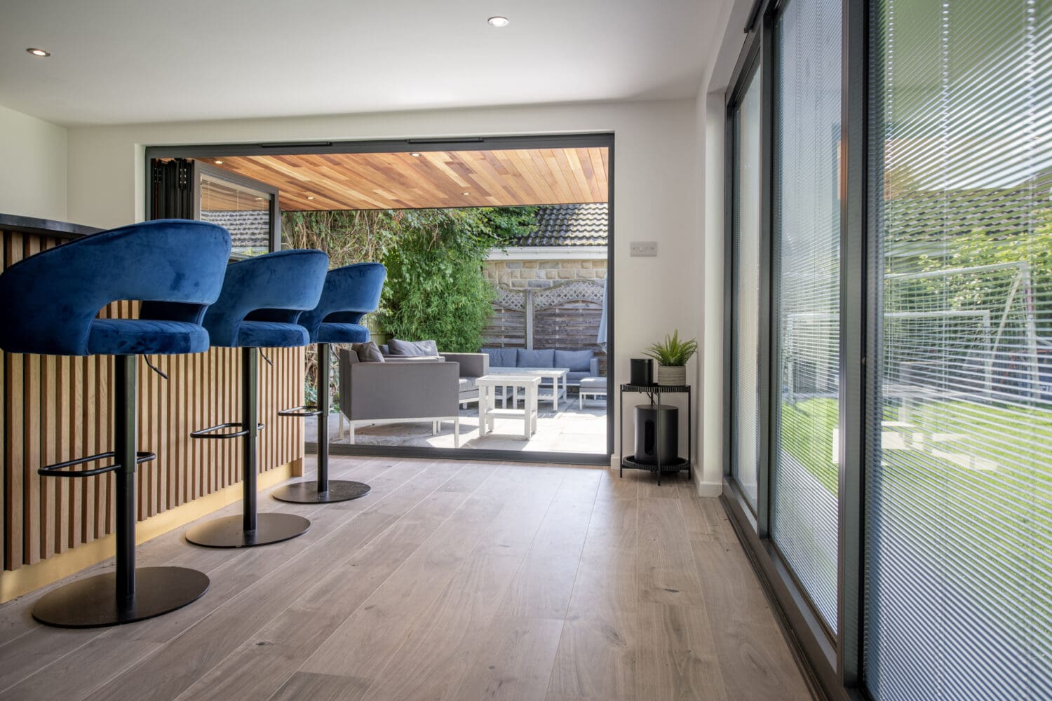Modern home interior with blue bar stools at a wooden counter, large sliding doors open to a patio with outdoor seating, and a green garden visible in the background. Natural light fills the space.