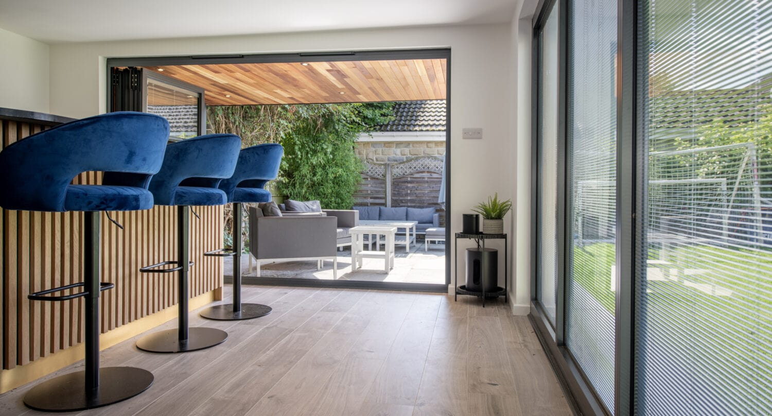 Modern home interior with blue bar stools at a wooden counter, large sliding doors open to a patio with outdoor seating, and a green garden visible in the background. Natural light fills the space.