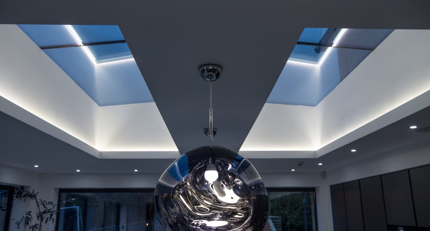 A modern pendant light hangs from the ceiling of a contemporary room featuring bifold doors, two large skylights, and recessed LED lighting, with large windows and a plant visible in the background.