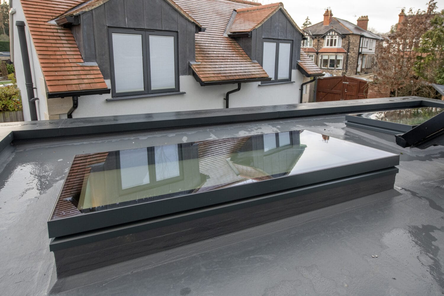 A modern flat roof with bespoke glass solutions features a large rectangular skylight on an extension of a house. In the background, older houses with pitched roofs, multiple windows, and distinct front doors are visible.