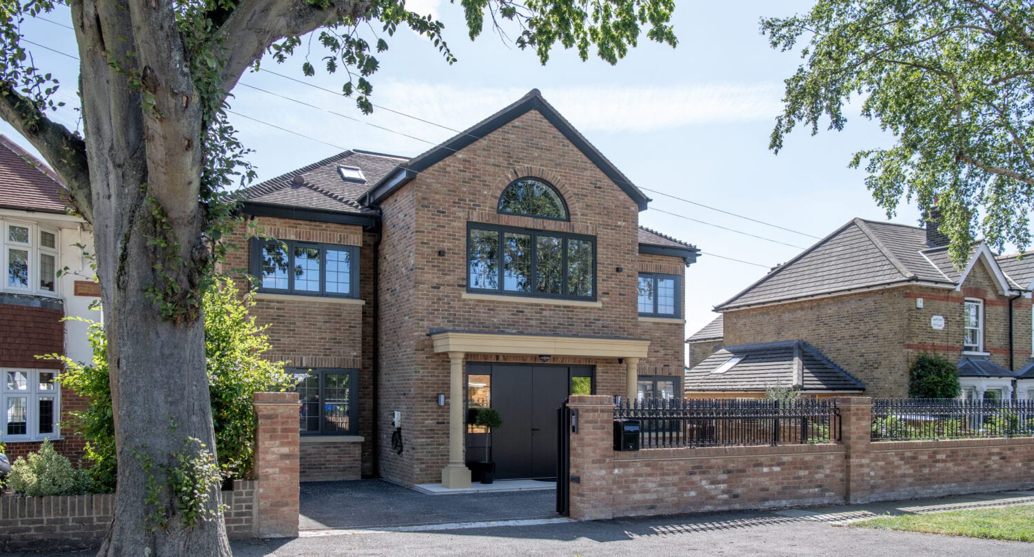 A modern two-story brick house with large windows, sleek sliding doors, a black double garage door, and a gated driveway, surrounded by a brick wall and trees on a sunny day.