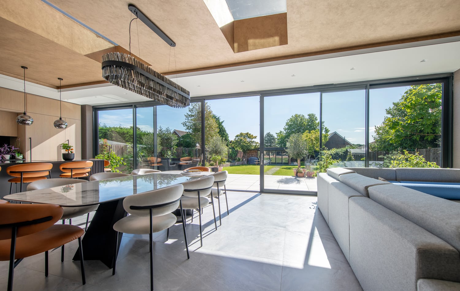 Modern dining and living area with large windows and bifold doors, letting in sunlight and offering a view of a green garden. The room features a long dining table, contemporary chairs, and a sectional sofa.