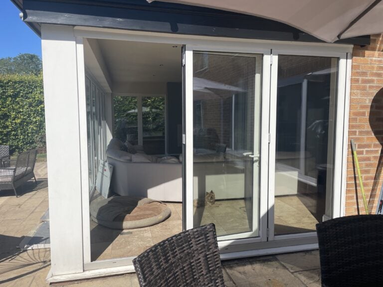 A glass patio door, crafted with bespoke glass solutions, opens onto a sunlit paved area with outdoor chairs. Inside, a sofa and a dog bed are visible while a small cat sits behind the glass, gazing outside at green hedges and brick walls.
