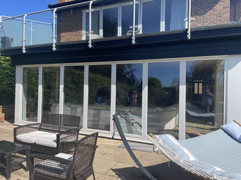 Patio area with wicker chairs, a table, and a hammock in front of a modern house featuring bespoke glass solutions like large sliding bifold doors and a glass balcony above, reflecting the outdoor scene and the person taking the photo.