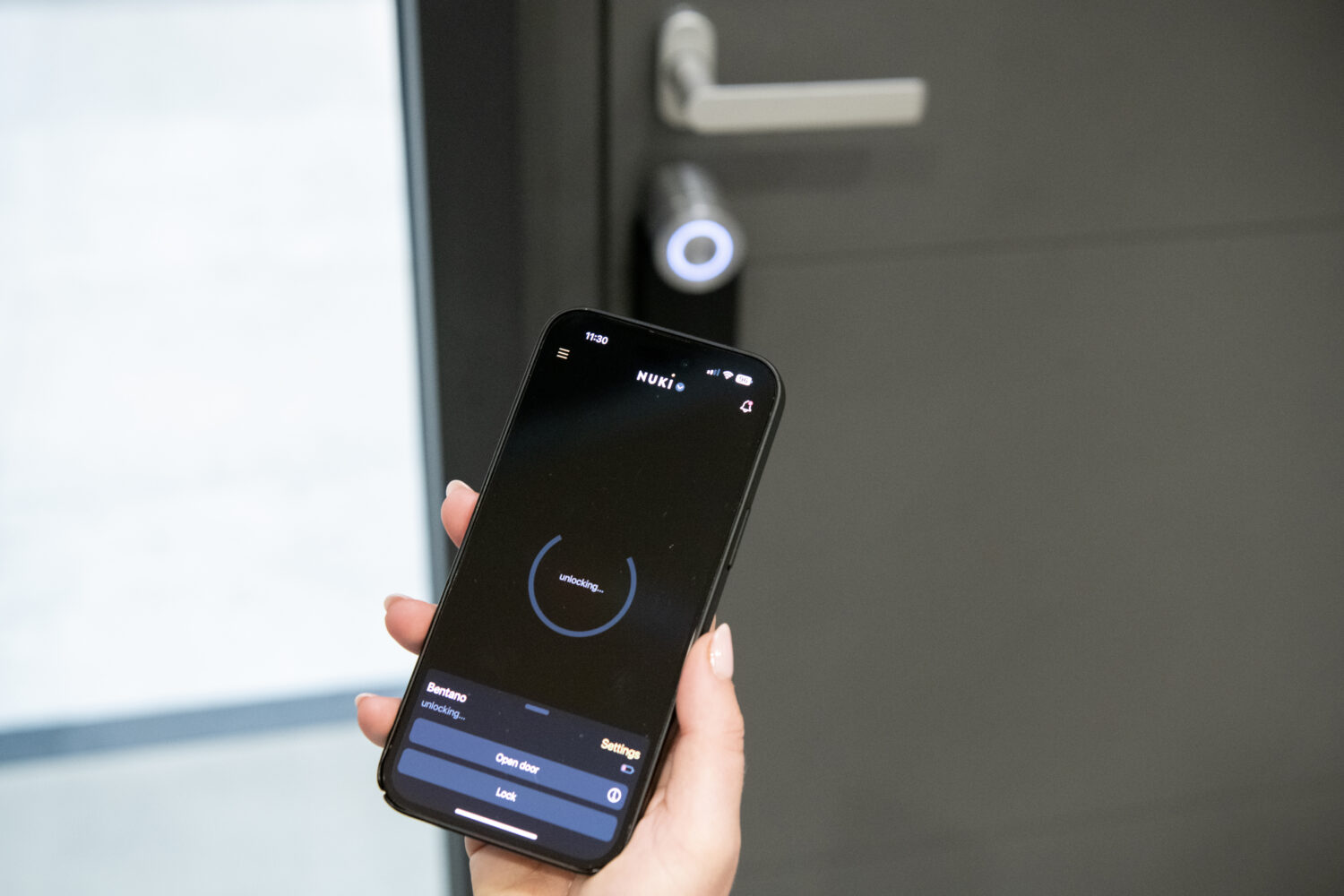 A person holds a smartphone with a smart lock app open, unlocking a modern electronic lock on sleek bifold doors in a minimalistic setting.