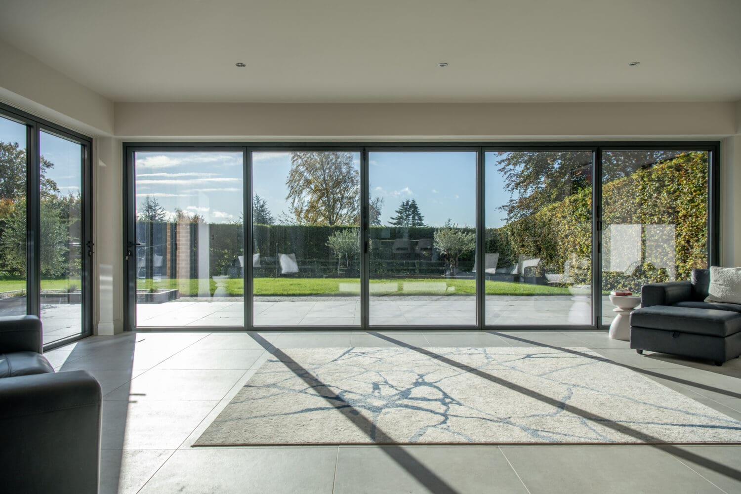 A modern living room with large floor-to-ceiling sliding doors opens to a garden. Sunlight fills the space, which features a marble-patterned rug and black sofas on tiled flooring, complemented by bespoke glass solutions for a sleek finish.