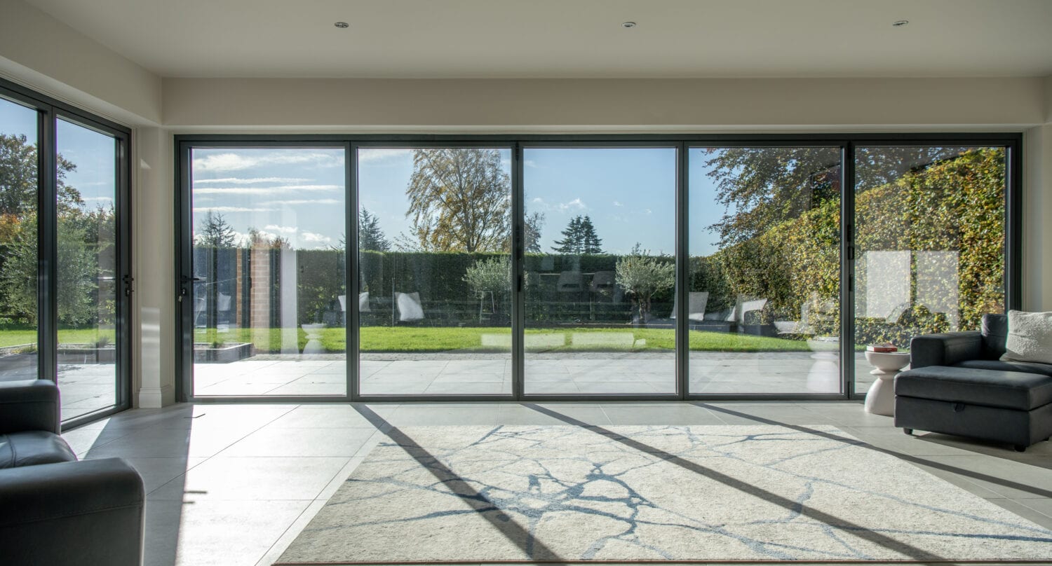 A modern living room with large floor-to-ceiling sliding doors opens to a garden. Sunlight fills the space, which features a marble-patterned rug and black sofas on tiled flooring, complemented by bespoke glass solutions for a sleek finish.