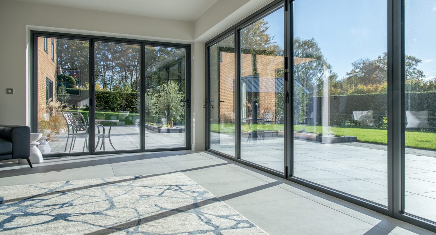 Bright modern room with large floor-to-ceiling sliding doors opening to a sunny patio and green garden, featuring a patterned rug and outdoor furniture.