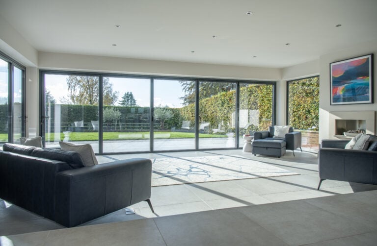 Modern living room with large floor-to-ceiling sliding doors overlooking a garden. The room features dark sofas, a rug, an armchair, and wall art above a fireplace. Sunlight from the bespoke glass solutions fills the bright, open space.
