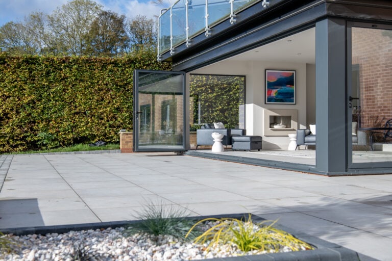 Modern house with large bifold doors open to a spacious patio. Inside, a light gray sofa, white stool, fireplace, and abstract wall art add style. The tiled patio features a rock and plant garden in the foreground.