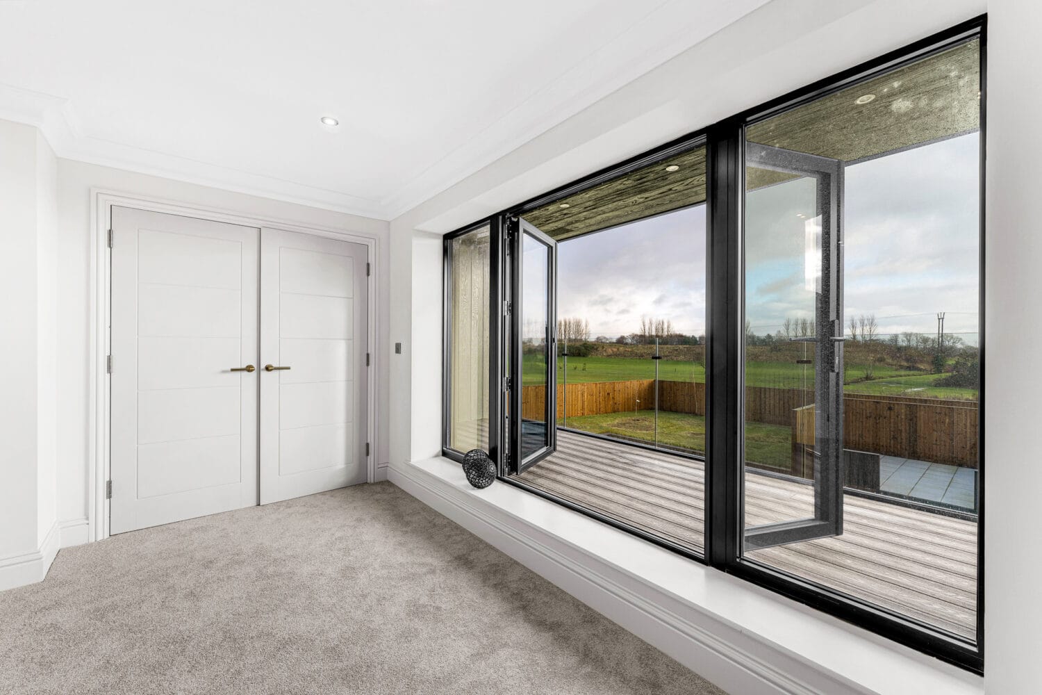A bright, modern room with light carpet, white double doors, and large black-framed sliding doors opens to a wooden deck. The windows overlook a fenced green field and trees under a cloudy sky.