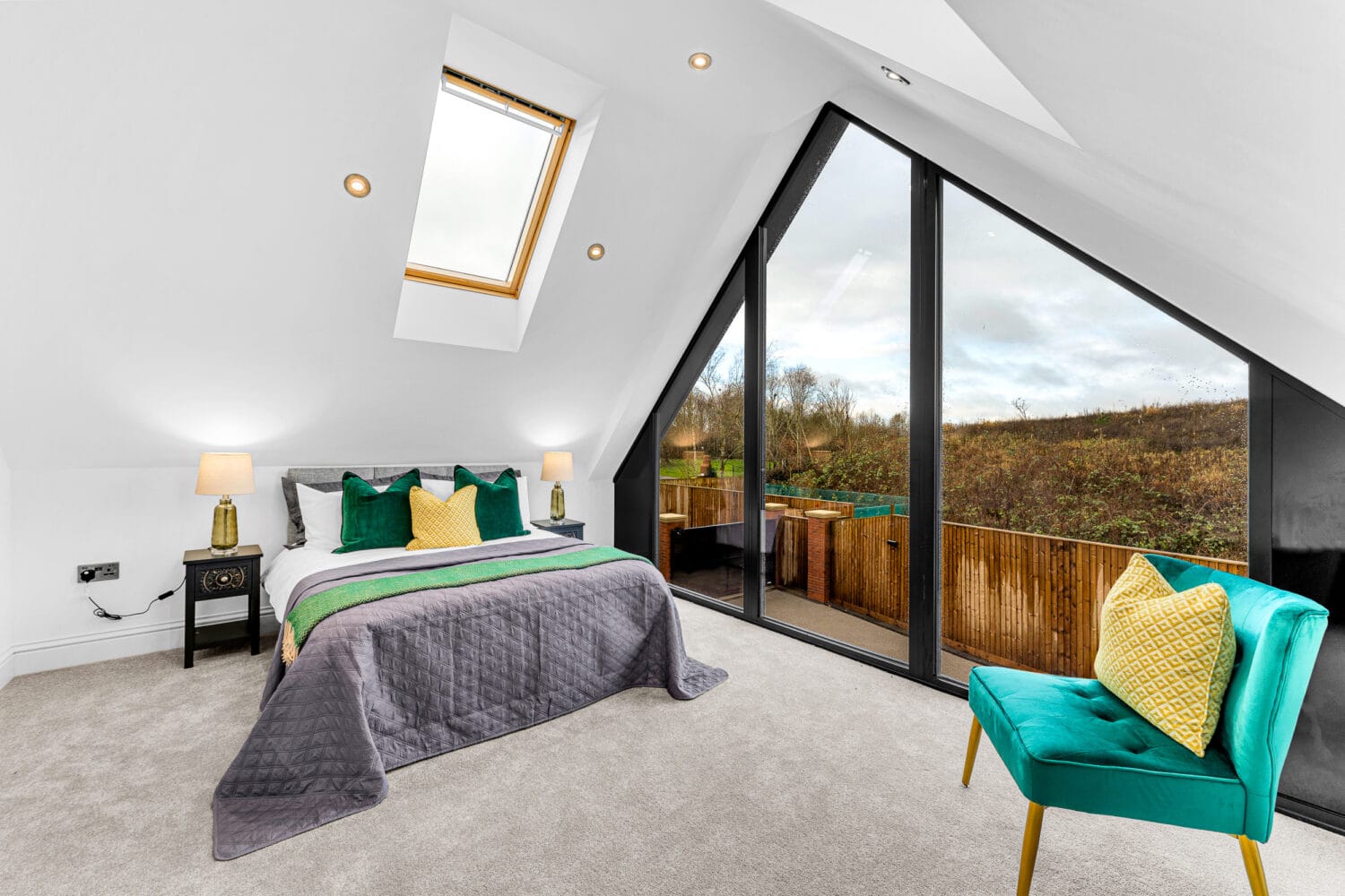 A modern attic bedroom with a large floor-to-ceiling triangular window, skylight, double bed with green and yellow pillows, teal chair, bespoke glass solutions, bedside table, and neutral carpet overlooking a scenic outdoor view.
