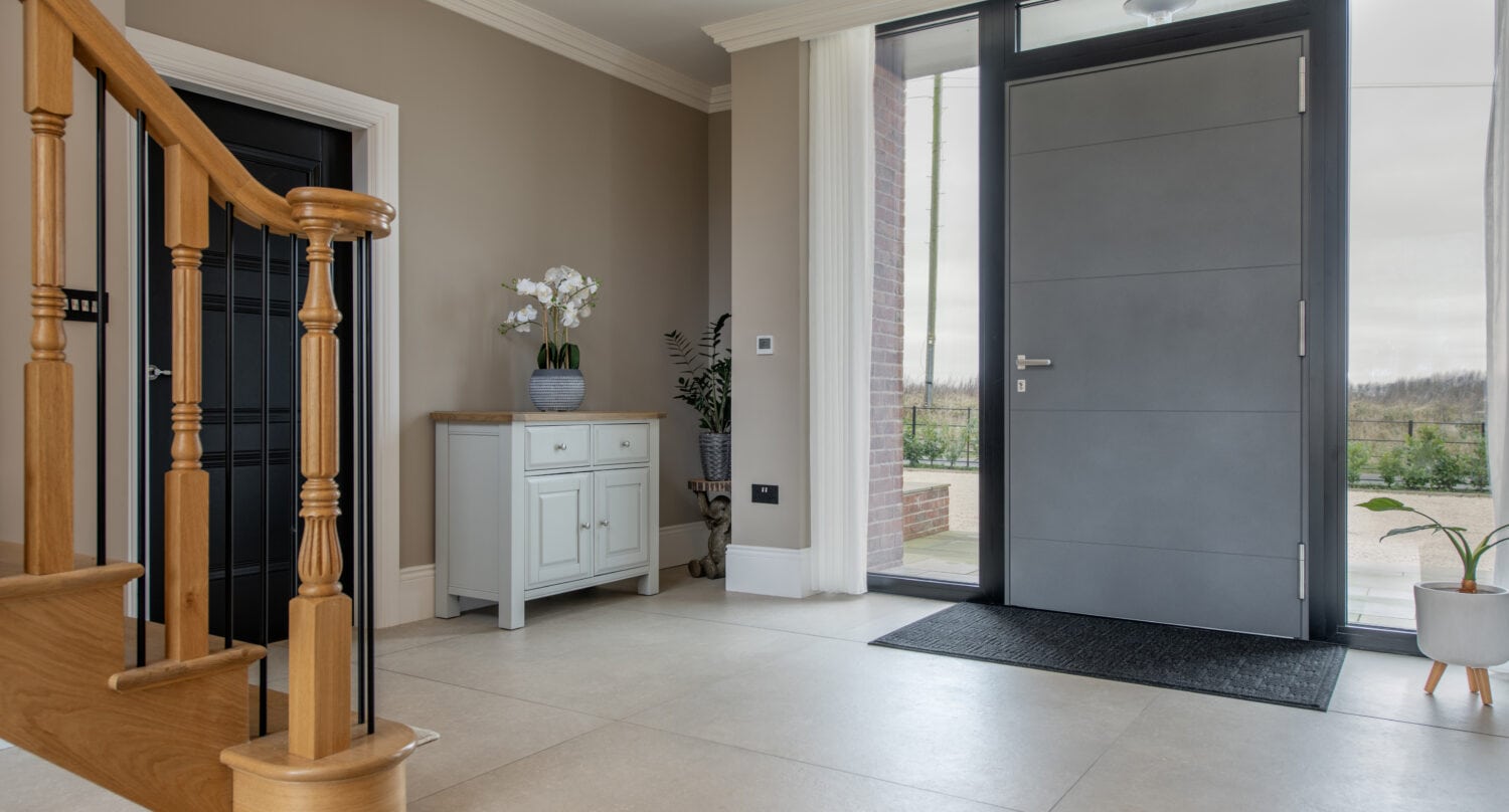 Modern entryway with a gray front door, large windows, light tile flooring, a wooden staircase, a white cabinet with potted orchids, and outdoor scenery visible through glass bifold doors.